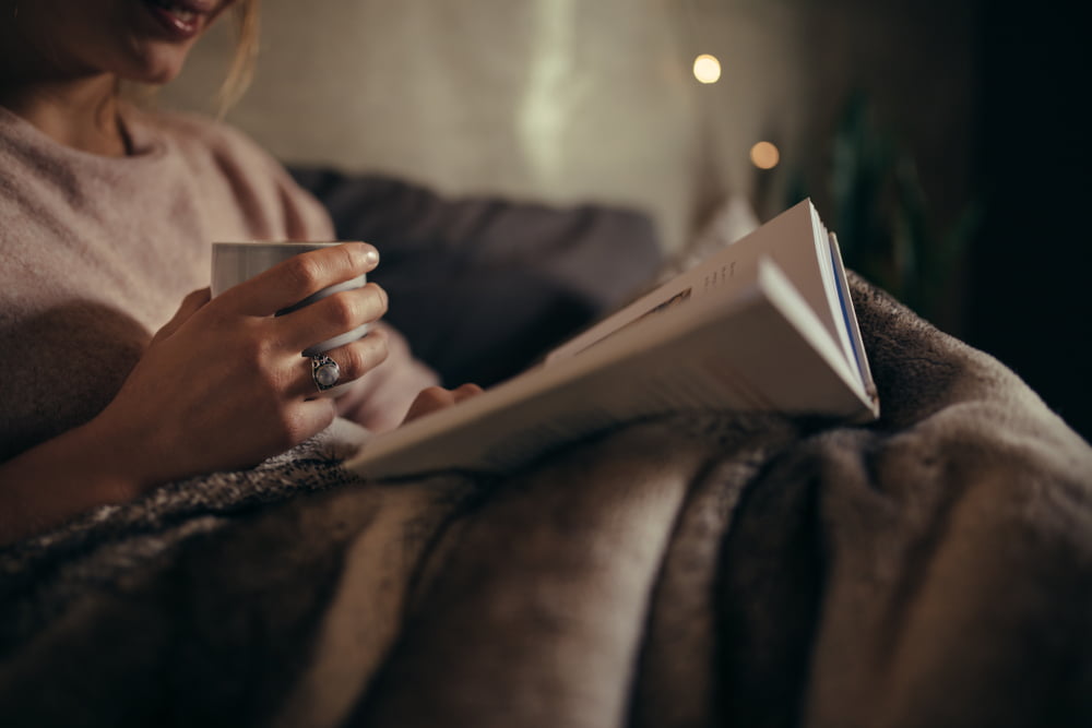 Cropped shot of female hands with book and coffee. Female reading book on bed at night.